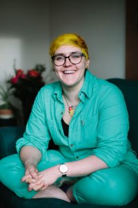 This is a picture of Fiona, a white woman with bright yellow hair. She is smiling and sitting in a relaxed cross legged position, wearing a turquoise suit.