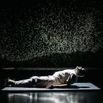 Performer Ramesh Meyyappan in a white shirt and grey trousers. Ramesh is lying on a black mat, with his head and torso lifted forwards and his legs on the floor. His left arm is beside him on the floor with fingers stretched out. The back wall is dark with thousands of tiny white lights projected. Photo taken by Mihaela Bodlovic.