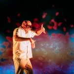 Performer Ramesh Meyyappan in a white shirt and grey trousers. Ramesh is stood up sideways with his left arm out blocking his eyes and his right arm out to the side with fingers pointing down. His mouth is wide open. The background is projected with a red and orange explosion. Photo taken by Mihaela Bodlovic.