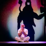 Performer Ramesh Meyyappan in a white shirt and grey trousers. Ramesh is sitting facing forward, crossed leg on a black mat with his palms together beneath his chin. The back wall has a projection of a large shadow shape with a ball of colours surrounding it. Photo taken by Mihaela Bodlovic.