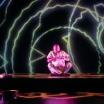 Performer Ramesh Meyyappan in a white shirt and grey trousers. Ramesh is sitting facing forward, crossed leg on platform with his hands creating two circles in front of his mouth and his lips are pursed. On the right edge of the platform are folded clothes and on the floor in front is a silver bowl. The back wall and floor has projections of various yellow and white circle outlines with jagged, wavy lines. Photo taken by Mihaela Bodlovic.