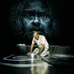 Performer Ramesh Meyyappan in a white shirt and grey trousers. Ramesh is kneeling on the floor holding on to and facing a silver bowl on the floor. There is a low, long platform at the back with folded clothes on the edge in the background. The back wall has a projection of a large face reflected in water. The floor has a white spiral projected onto it. Photo taken by Mihaela Bodlovic.